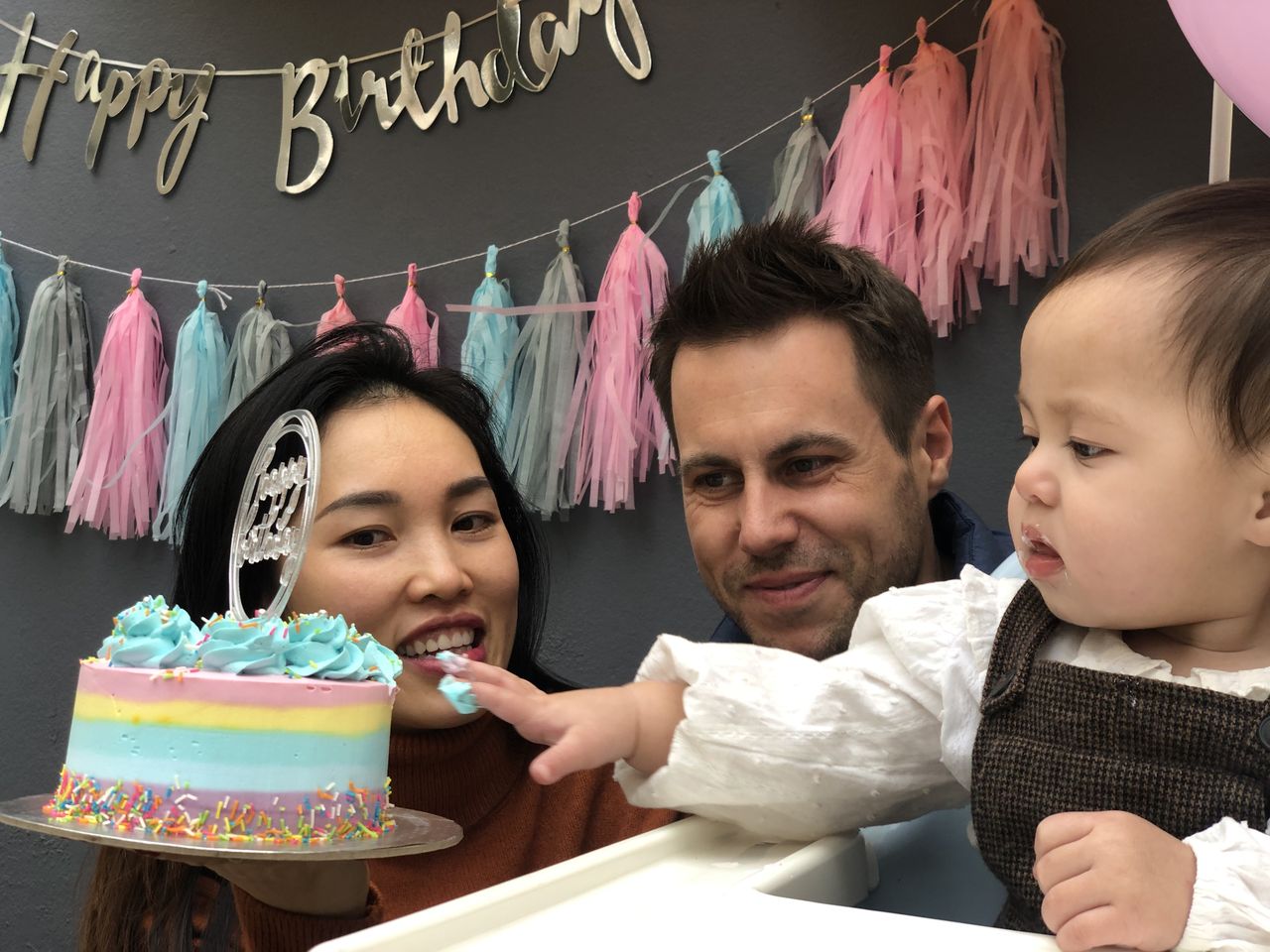 A guest couple with baby Lena reaching for a rainbow birthday cake under a Happy Birthday banner
