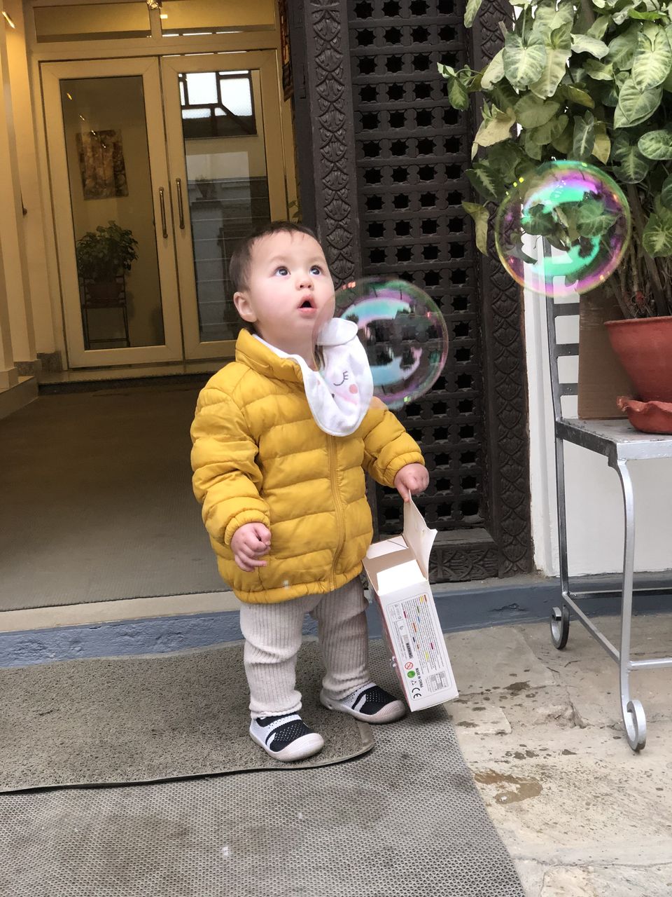 Toddler Lena in a yellow coat reaching for a soap bubble at the residence entrance